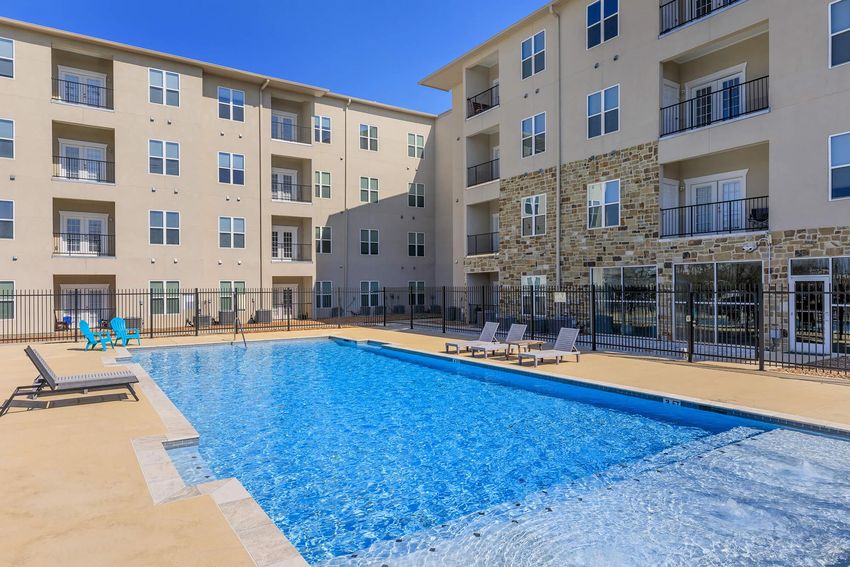 A large outdoor swimming pool surrounded by lounge chairs, with an apartment building in the background. The area is well-maintained, featuring a mix of stone and modern architecture, and includes a fenced enclosure for added privacy. Bright blue sky overhead.