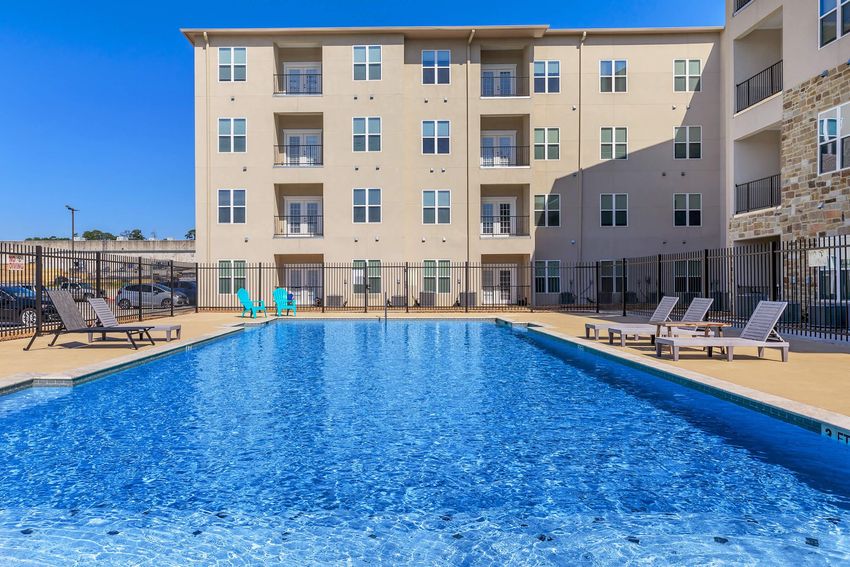 An outdoor swimming pool with crystal-clear water, surrounded by lounge chairs and a black metal fence. In the background, a multi-story apartment building features numerous windows and balconies under a clear blue sky.