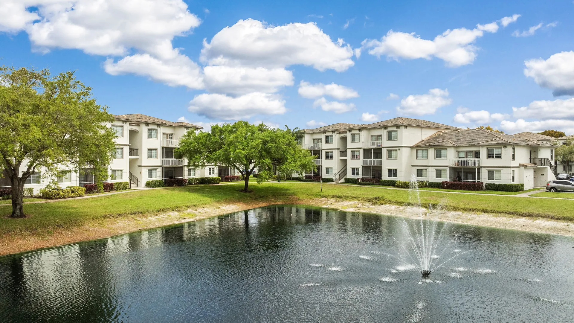 A serene view of an apartment complex by a pond with a fountain under a blue sky.