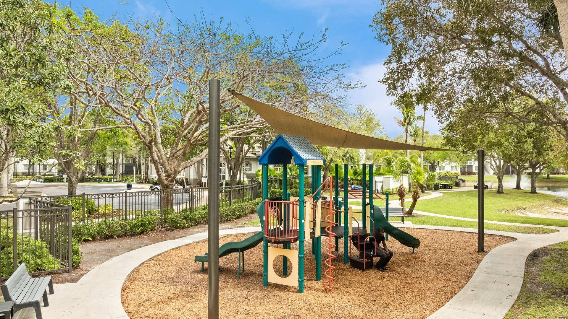 A colorful playground with slides and climbing structures under a shade sail, surrounded by green trees.