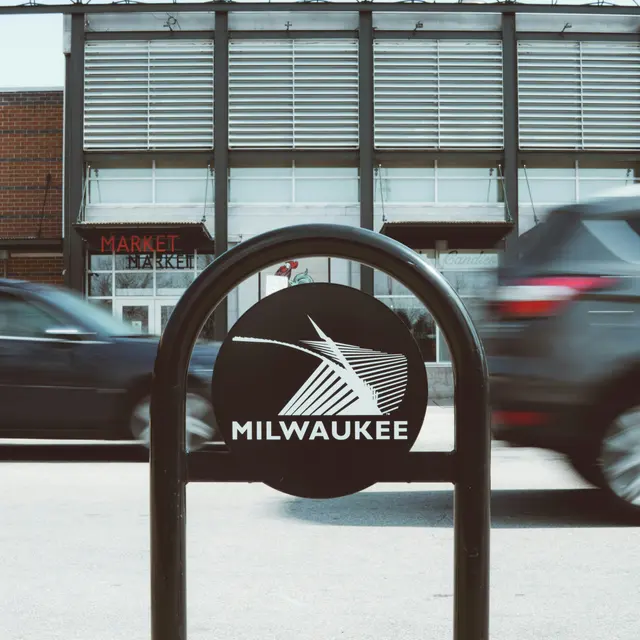 View of a street sign for Milwaukee with the Milwaukee Public Market in the background, featuring large letters that read 'MILWAUKEE PUBLIC MARKET'. Cars are passing by in front of the sign.
