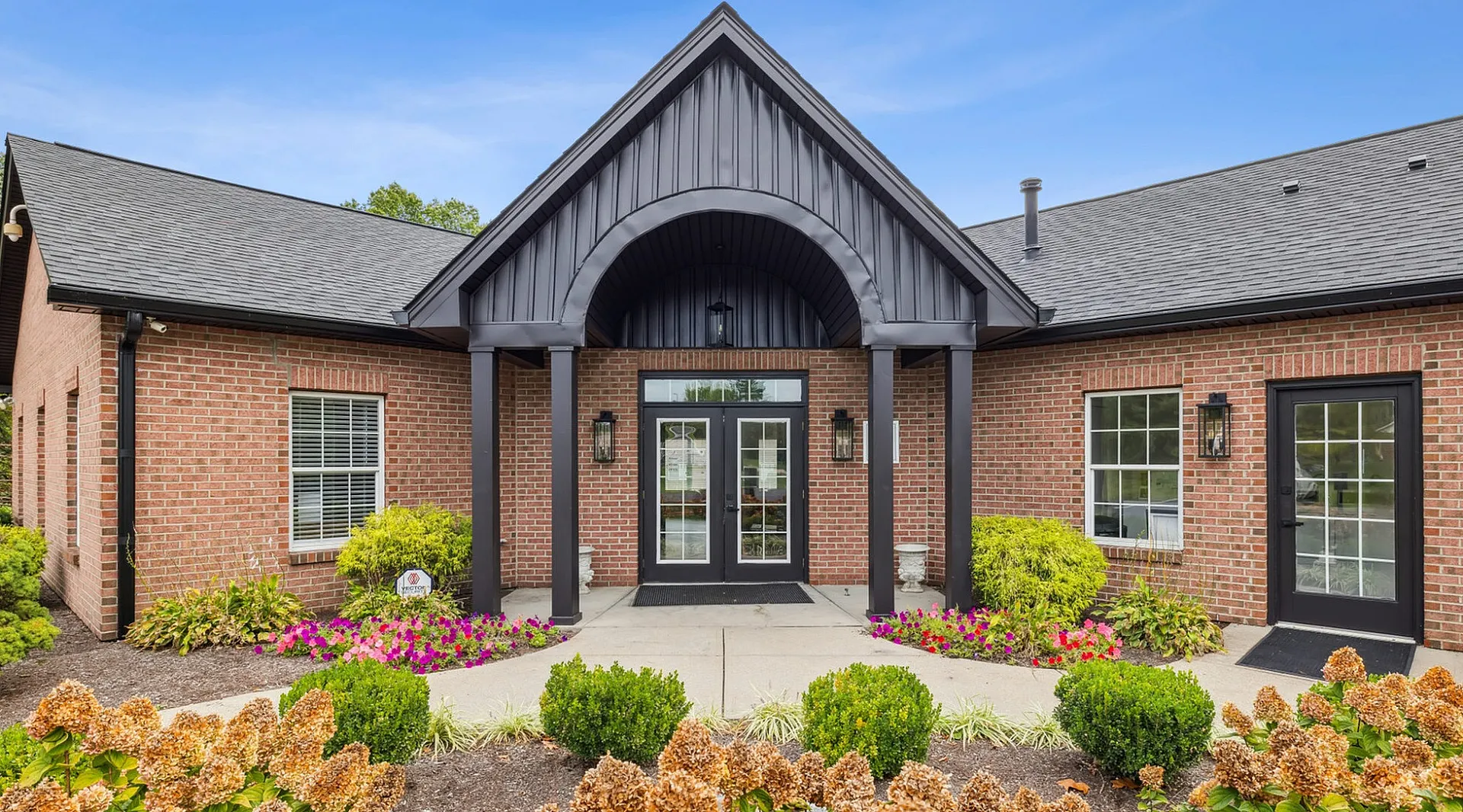 Charming brick building entrance with elegant double doors framed by lush landscaping and vibrant flower beds.