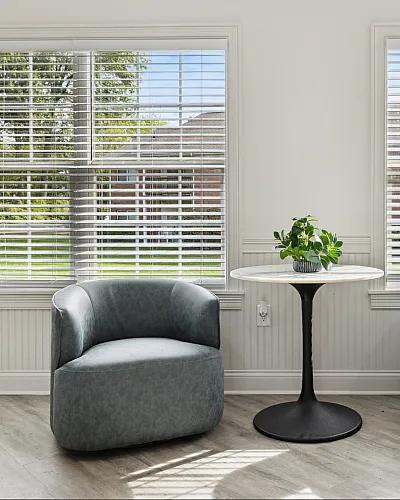 Bright sitting area featuring two modern grey armchairs, a round table with a lush plant, and large windows with views of a vibrant garden.