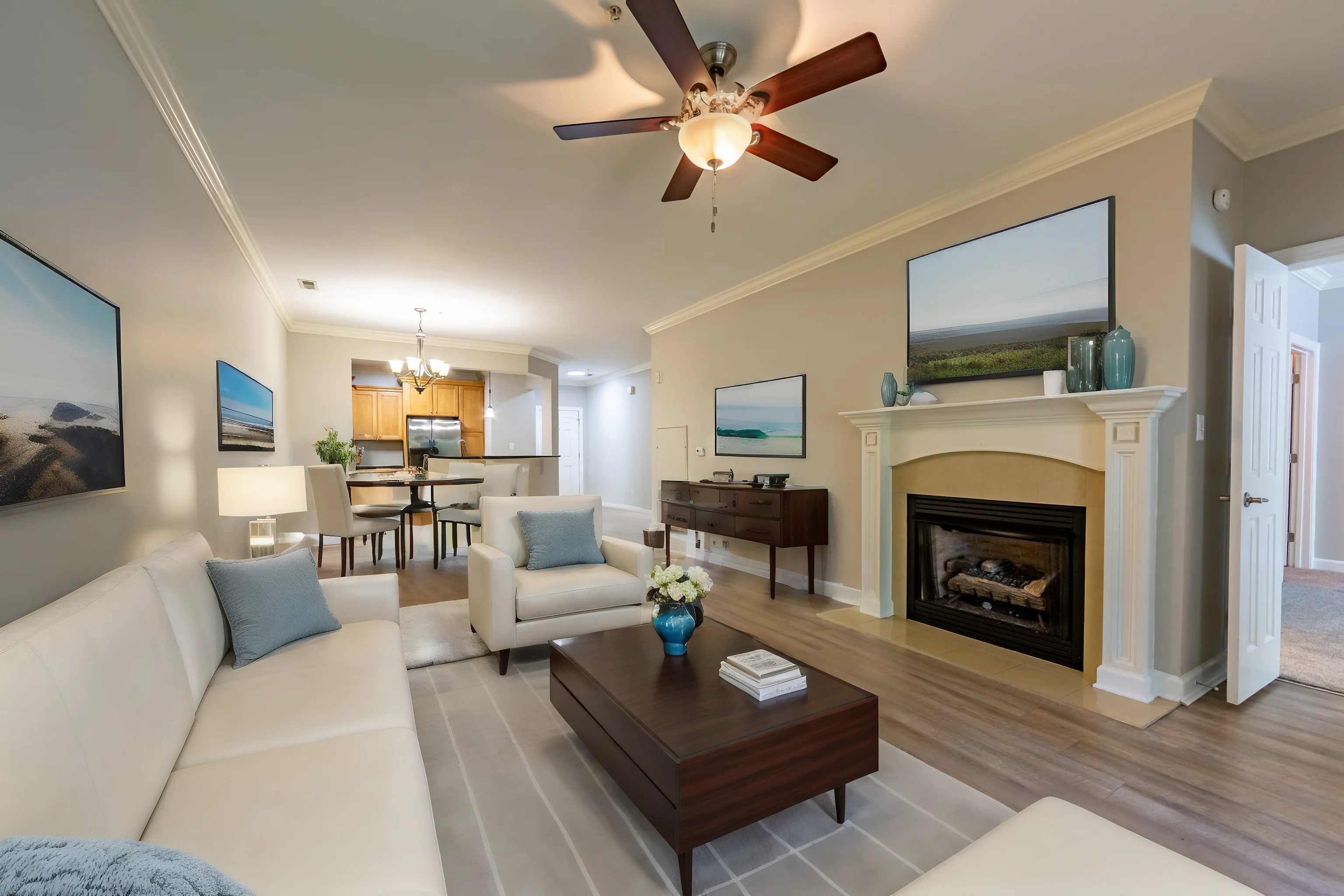 A modern living room featuring a white sectional sofa, a coffee table, and a fireplace. There are two framed images on the wall and a dining area visible in the background.