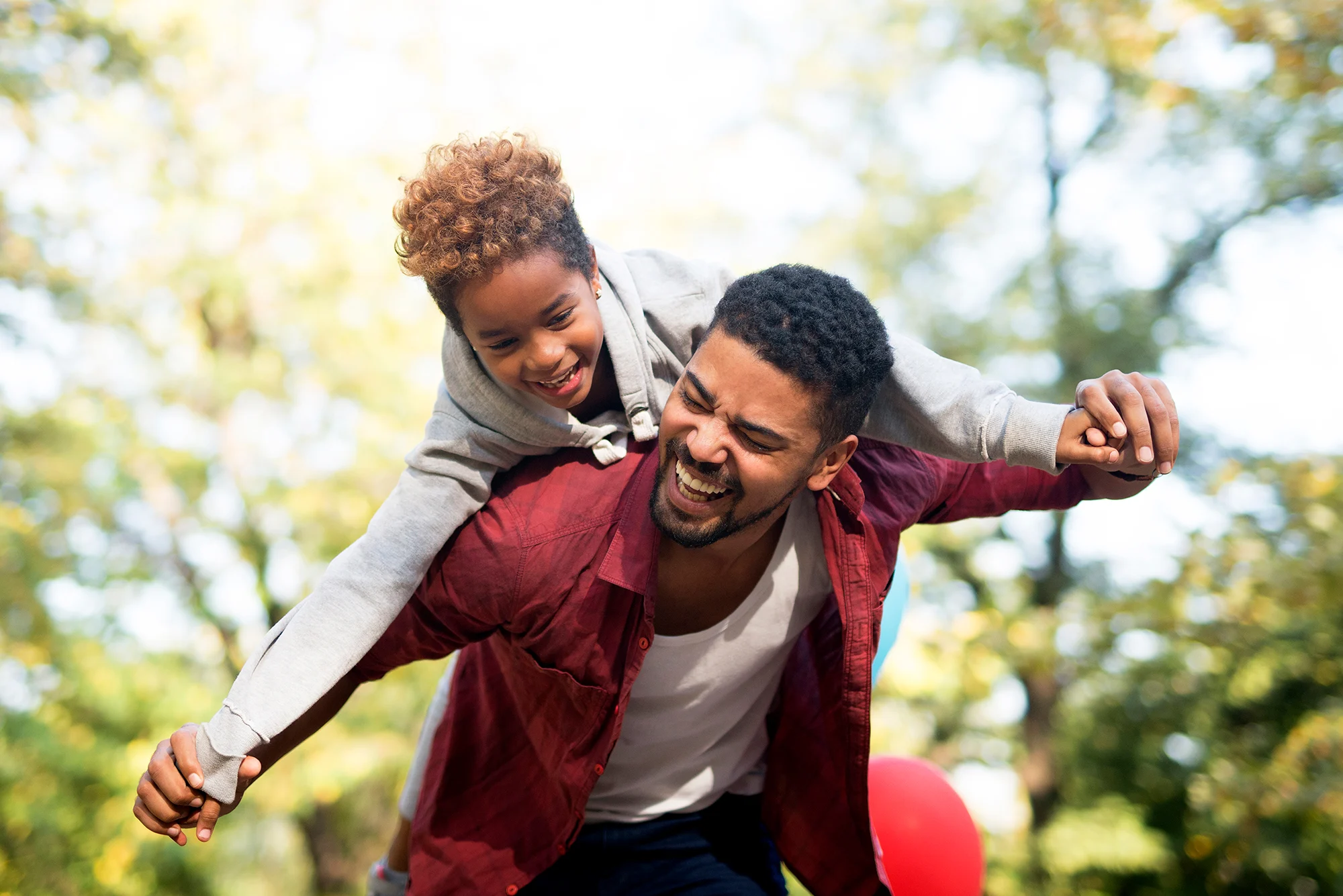 A joyful father carrying his son on his back in a park during a sunny day. The son is laughing while the father smiles broadly.