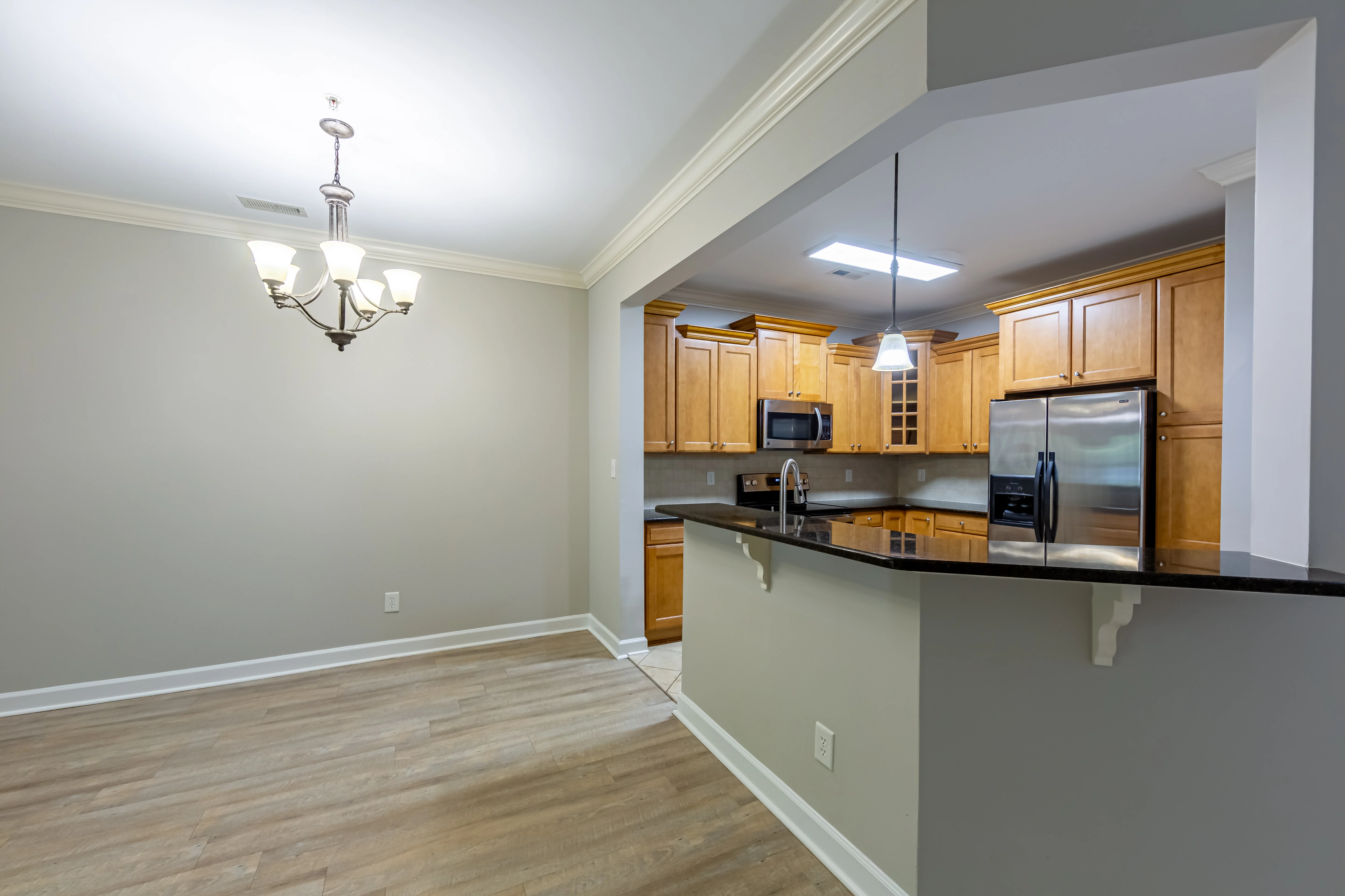 An open living space featuring a kitchen area with wooden cabinets, stainless steel appliances, and a dark countertop. There is a dining area with a chandelier and light-colored walls and flooring.