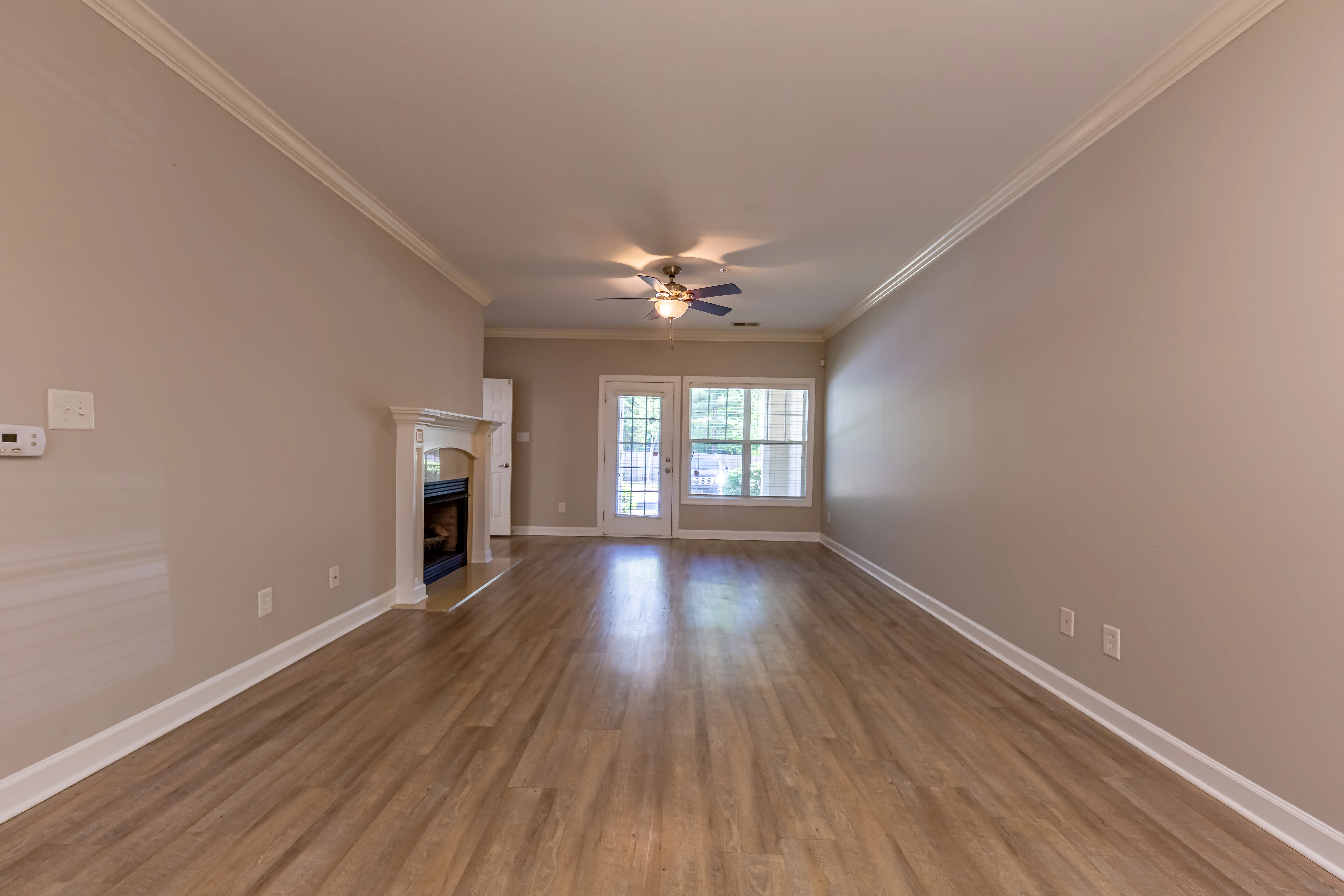 Spacious living room with light wooden flooring, a ceiling fan, and a fireplace on the left. Large windows allowing plenty of natural light.