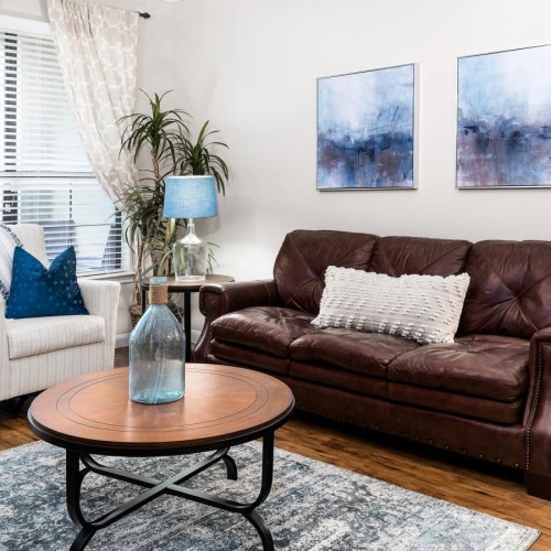 Apartments Near Space Center of Houston, TX - Ranch at City Park - Bright Living Room With Leather Sofa, Accent Chair, Round Coffee Table, Blue Lamps, and Large Window.