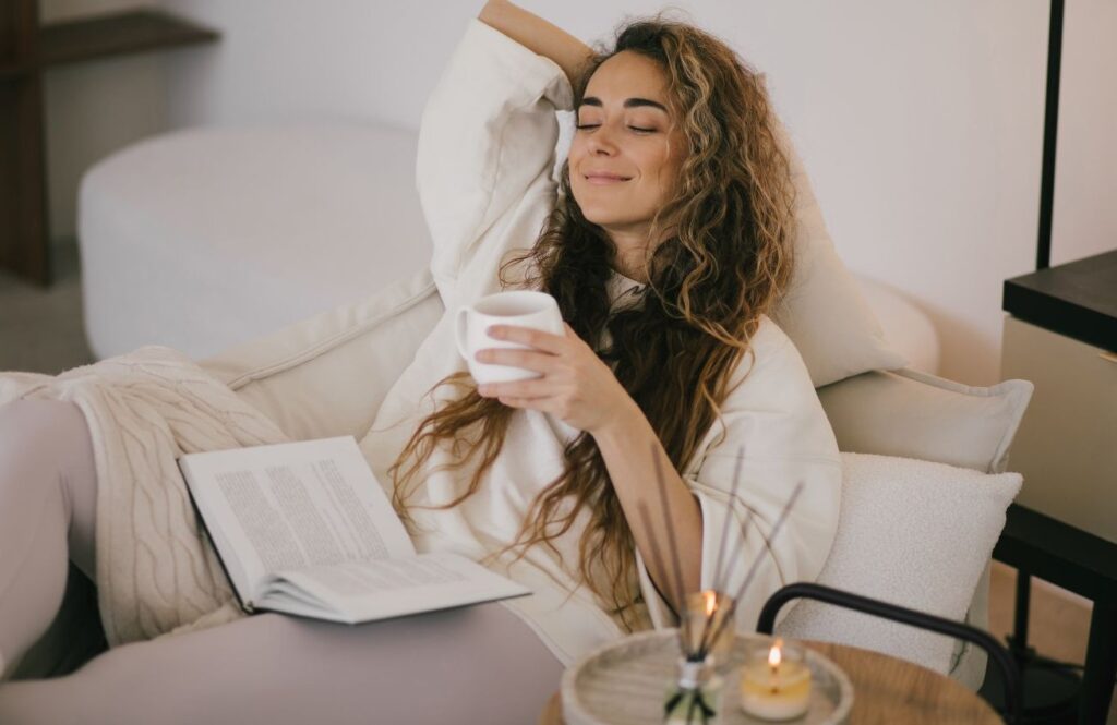 A smiling woman holding a white mug and an open book relaxing on a comfortable couch near burning candles.