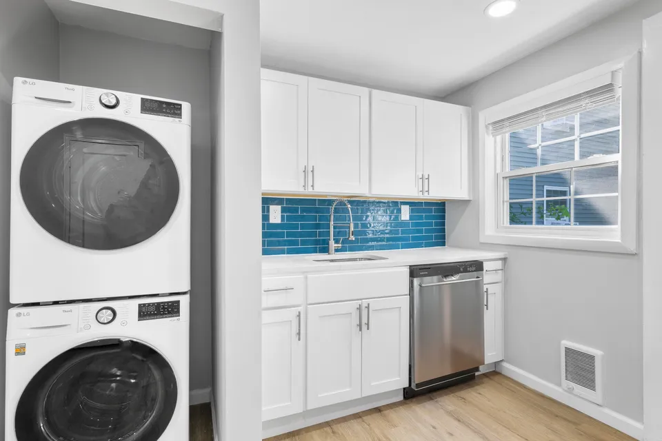 Modern kitchen with elegant white cabinetry, vibrant blue tiled backsplash, stainless steel dishwasher, and in-unit laundry for convenience.