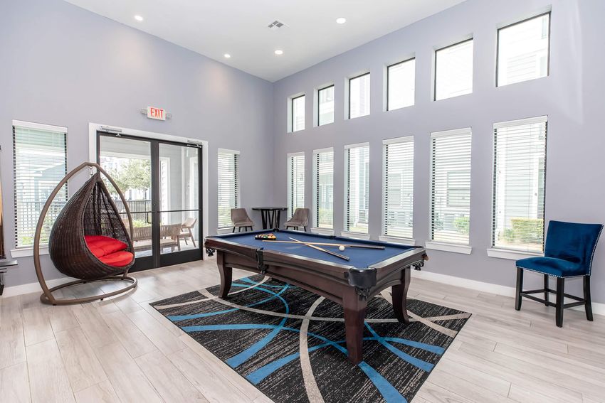 A modern indoor recreational area featuring a billiard table, a hanging circular chair with a red cushion, blue upholstered seating, large windows providing natural light, and a decorative rug that complements the room's design. The space is designed for relaxation and social interaction.