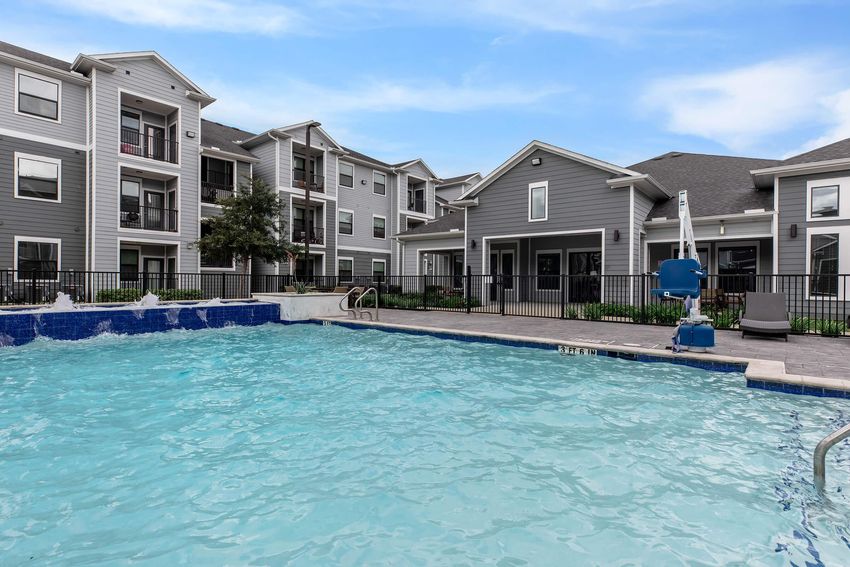 View of a swimming pool with clear blue water, surrounded by a patio area and apartment buildings in a residential complex. The pool features a ladder and a shallow area, with lounge chairs positioned around. Trees and landscaping add greenery to the setting, under a clear blue sky.