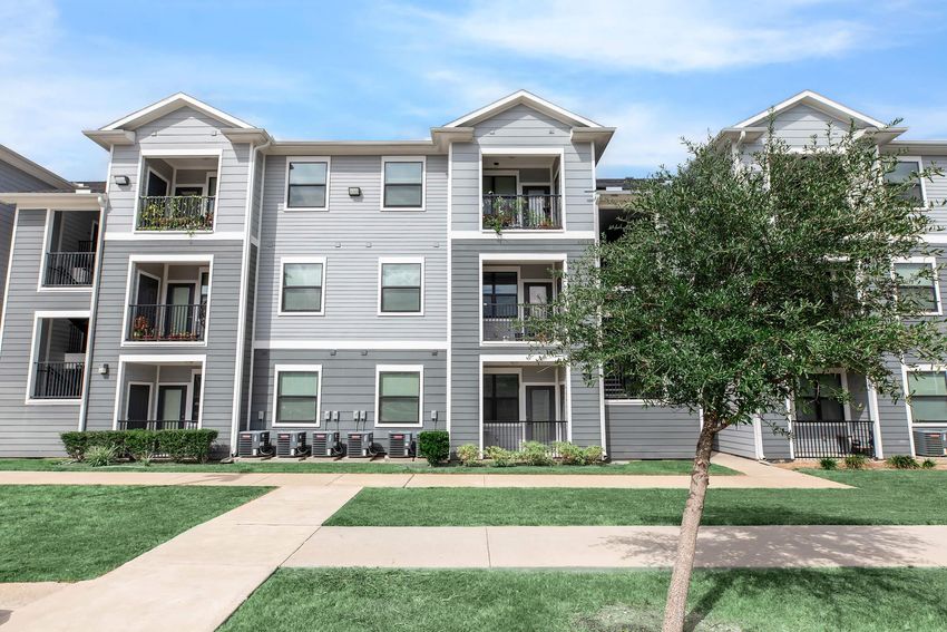 A modern apartment building with a light gray exterior featuring multiple balconies. Lush green landscaping surrounds the building, with a concrete pathway leading through the yard. Air conditioning units are visible outside each apartment, and a small tree is situated in the foreground.