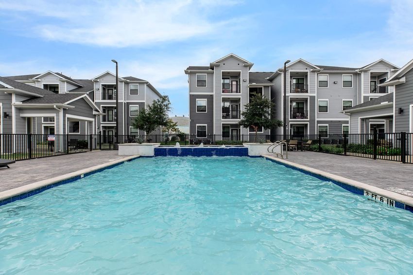 A clear swimming pool with light blue water, surrounded by a paved area. In the background, there are modern apartment buildings with gray siding and balconies. The scene is set under a bright blue sky, creating a welcoming atmosphere for residents and visitors.