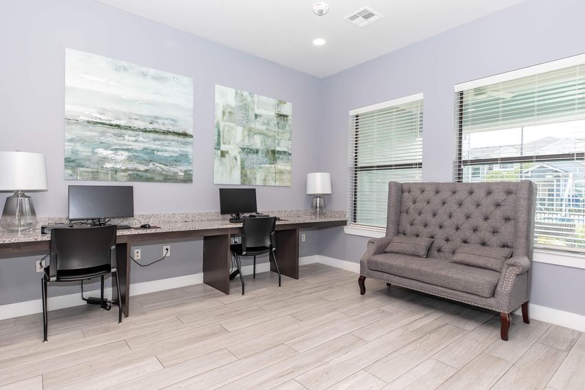 A modern office space featuring two computer desks with black chairs, a gray tufted sofa, and two abstract paintings on the walls. Natural light filters through window blinds, and the floor is covered in light-colored tiles, creating a bright and inviting atmosphere.