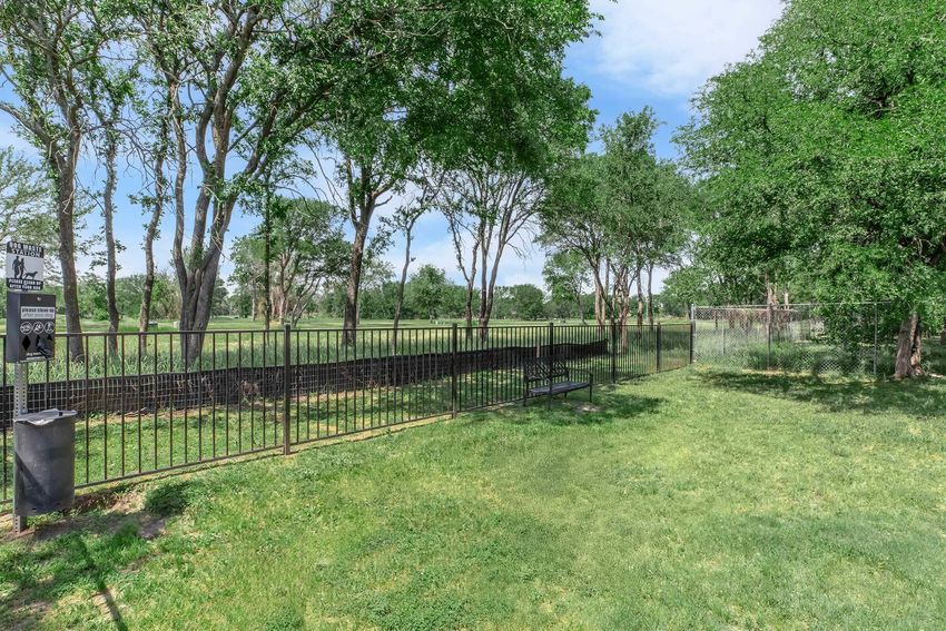A green park area featuring a black metal fence bordering a grassy field. Several trees provide shade, and a bench is positioned along the path. In the background, a playground structure can be seen, indicating a family-friendly environment. The sky is clear with a few clouds.