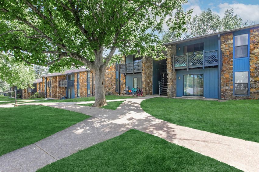 A well-maintained residential complex featuring two-story stone and wood units. Lush green lawns surround the buildings, and a large tree provides shade. A paved walkway intersects the grassy area, leading to the entrances of the apartments. The scene is bright and inviting, indicating a pleasant living environment.
