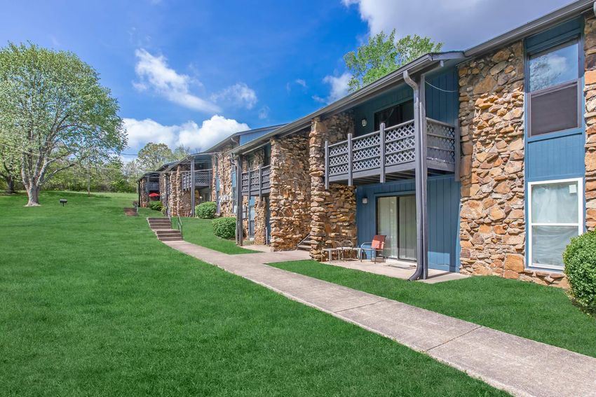A scenic view of a multi-unit stone apartment building set amidst lush green grass and trees under a blue sky with fluffy clouds. The pathway leads to the entrances of the apartments, and a few chairs are placed on the balconies, suggesting a welcoming outdoor space.