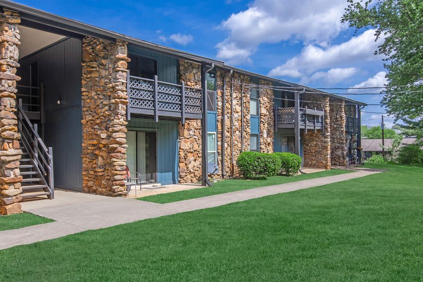 A view of a multi-unit apartment building featuring stone exterior walls and balconies. The building is flanked by green grass and a clear blue sky with fluffy clouds, and there are concrete pathways leading to the entrance on the ground level.