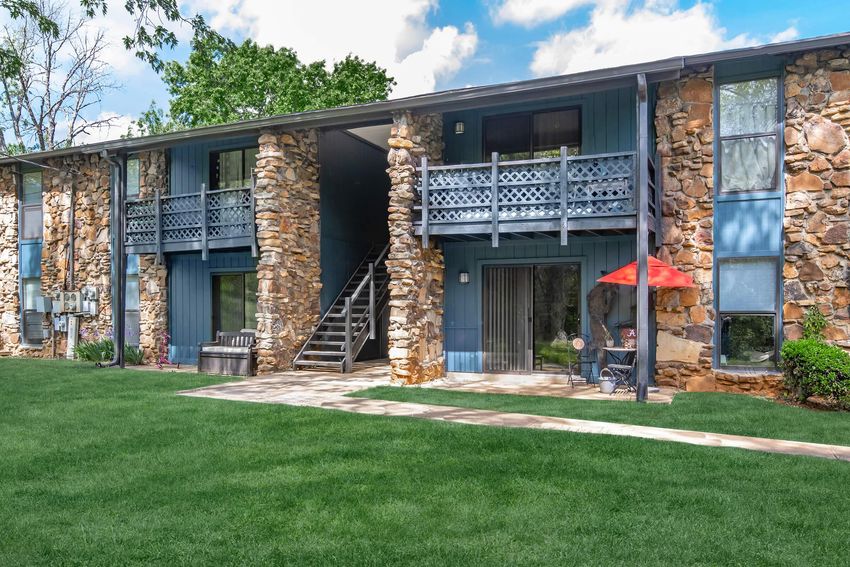 A two-story apartment building with stone and blue siding. The structure features balconies with railings, and a staircase leading to the upper level. In the foreground, there is a well-maintained green lawn, a sidewalk, and an outdoor seating area with a red umbrella. Bright blue sky with scattered clouds above.