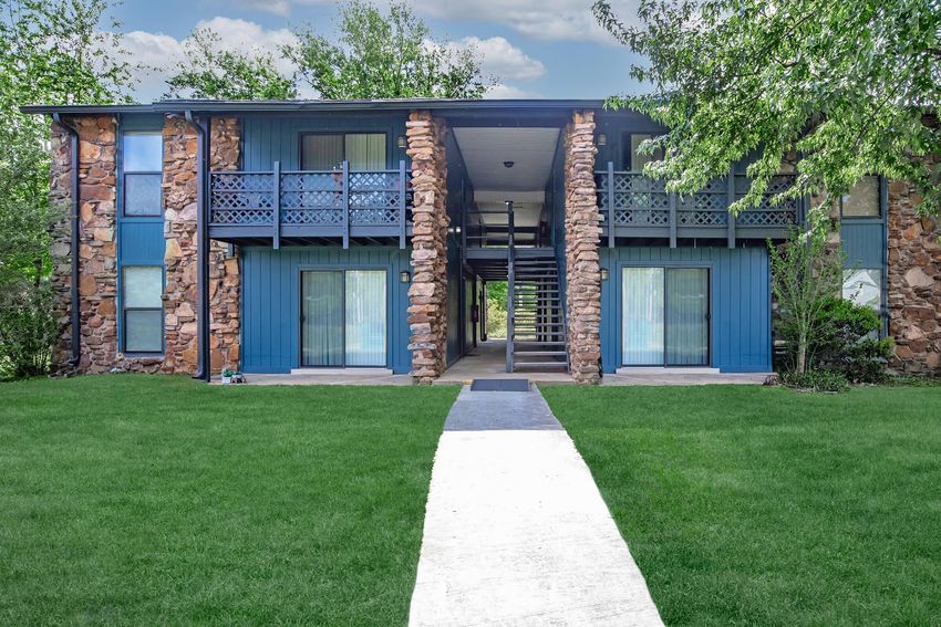 A two-story apartment building with stone accents and blue siding. The building features multiple balconies, large windows, and a well-maintained lawn in front. A concrete pathway leads to the entrance, surrounded by trees and greenery.