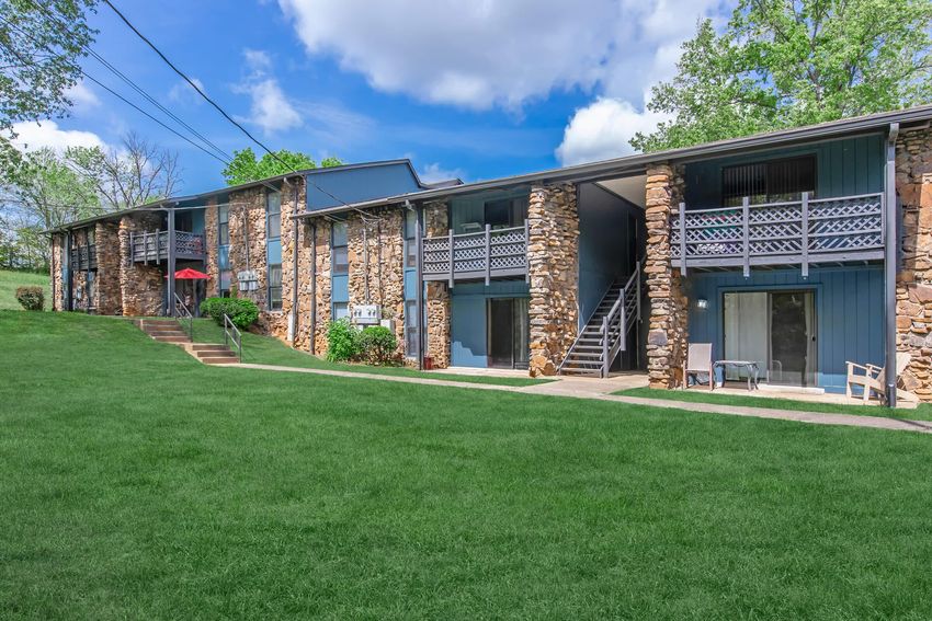 A two-story apartment building featuring stone and blue siding, surrounded by a green lawn. There are outdoor stairs, a small patio area with a table, and an umbrella. The sky is bright with clouds, and trees are visible in the background.