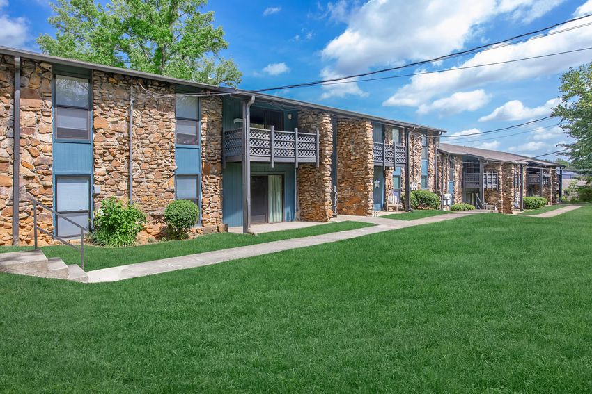 Two-story apartment building with a rustic stone facade, surrounded by well-maintained green lawns and small shrubs. The sky is partly cloudy with blue and white hues, and there are walkways leading to the entrance of the apartments. Power lines run overhead.