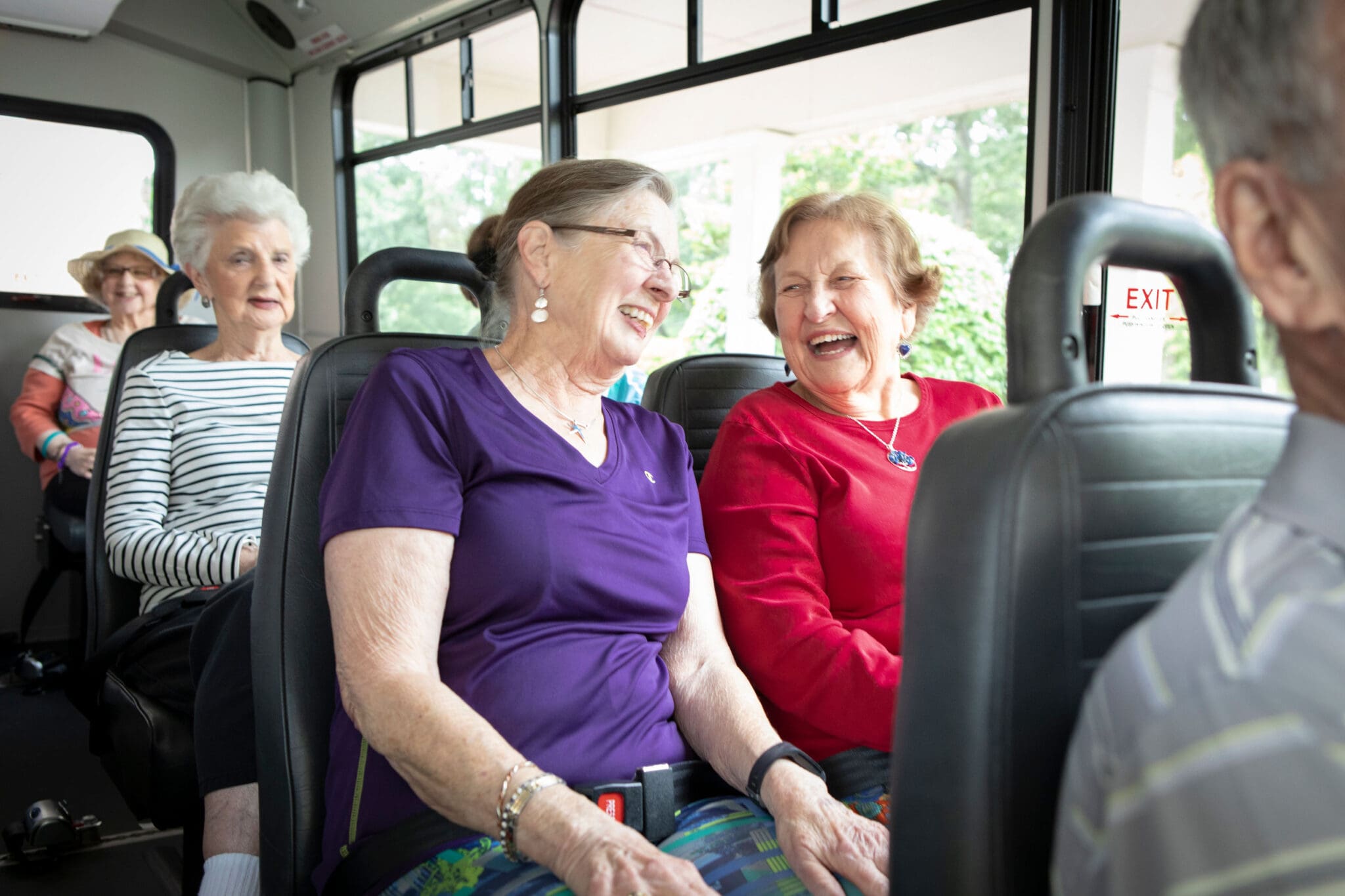 A tour bus/van is full of seniors heading into town. The image focuses on two women sitting next to each other laughing at each other. One is wearing a purple shirt and the other is wearing a red shirt. They have seatbelts on for safety.