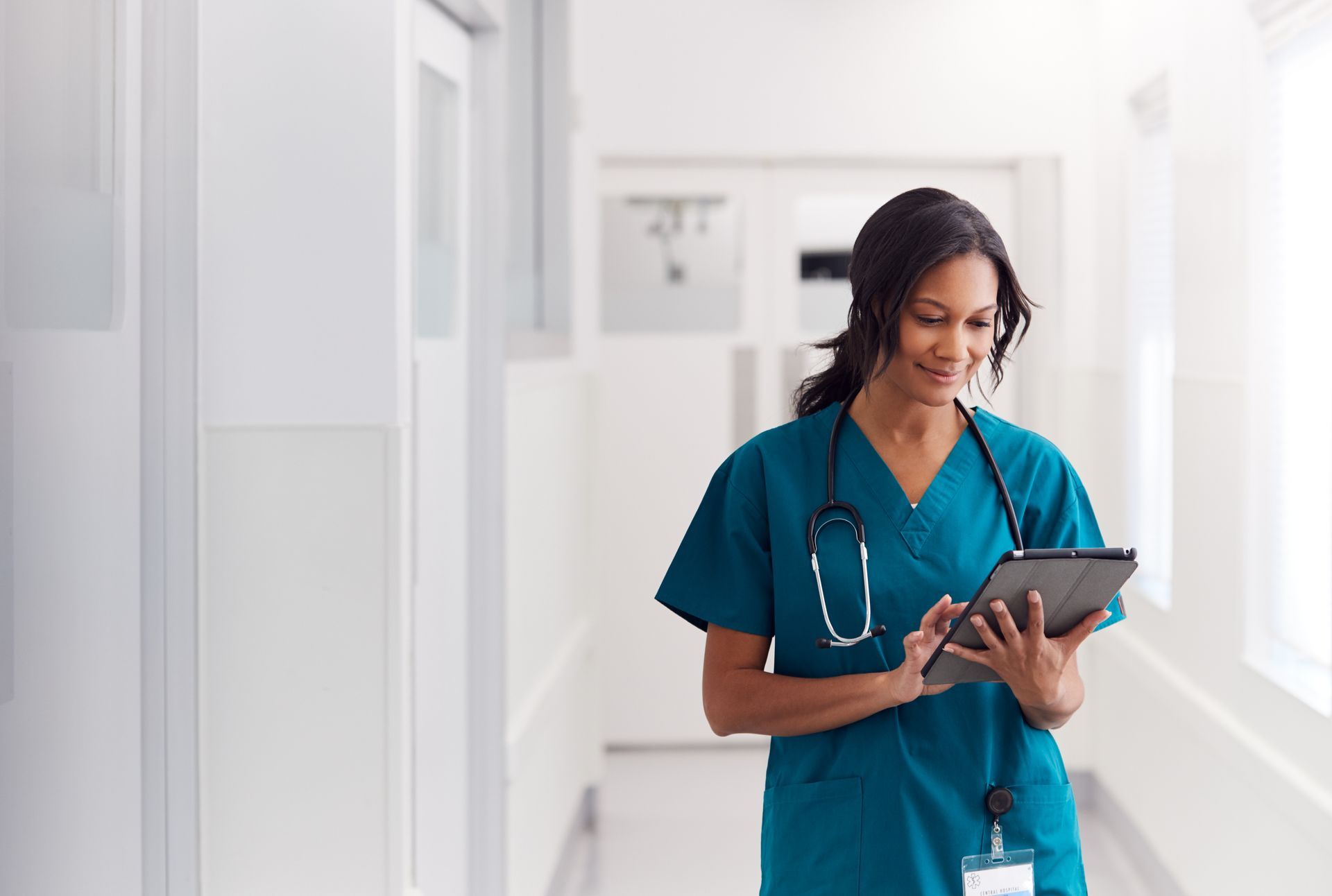 Healthcare worker in teal scrubs using a tablet in a bright hospital hallway.