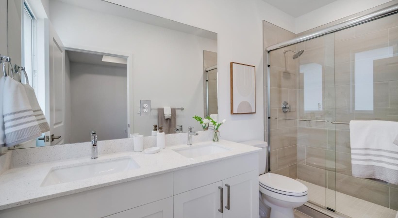 Palmetto Bay, FL, Townhomes with an Attached Garage - The Addison - A Bathroom with a Dual Vanity, White Cabinetry, Large Mirror, Empty Photo Frame, and a Framed Glass Shower Enclosure.