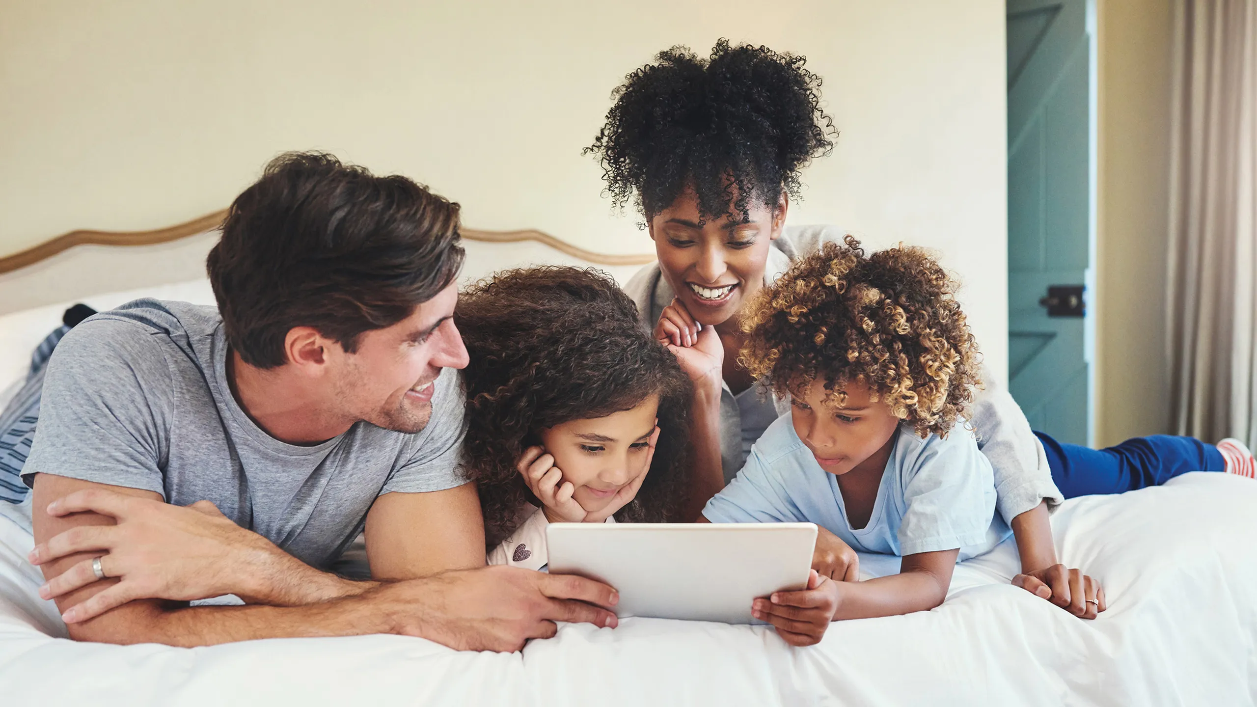 Happy family lying on a bed enjoying a tablet together in a cozy home setting.