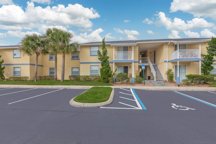 A two-story apartment building with a yellow exterior, featuring balconies and surrounded by lush greenery. In the foreground, there is a parking lot with marked spaces, including a blue and white handicapped parking area. The sky is bright with fluffy clouds.