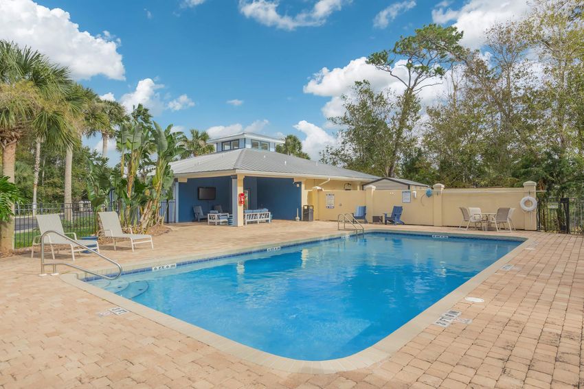 A bright outdoor pool area featuring a clear blue pool surrounded by brown pavers. There are several lounge chairs positioned next to the pool and a yellow building in the background with a gray roof. Lush green trees and palm plants are visible, under a partly cloudy sky.