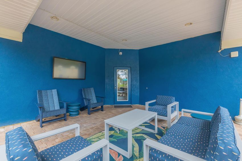 A cozy outdoor sitting area featuring four blue and white chairs and a white coffee table on a patterned rug. The walls are painted in a vibrant blue, and a television is mounted on the wall. A glass door adds light to the space, creating a comfortable setting for relaxation.
