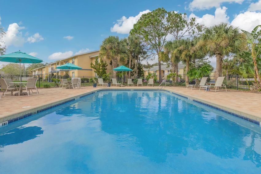 A bright, clear swimming pool surrounded by lounge chairs and umbrellas. Lush palm trees and tropical plants line the pool area, with a sunny sky filled with fluffy white clouds in the background. The pool is well-maintained, inviting relaxation and leisure.