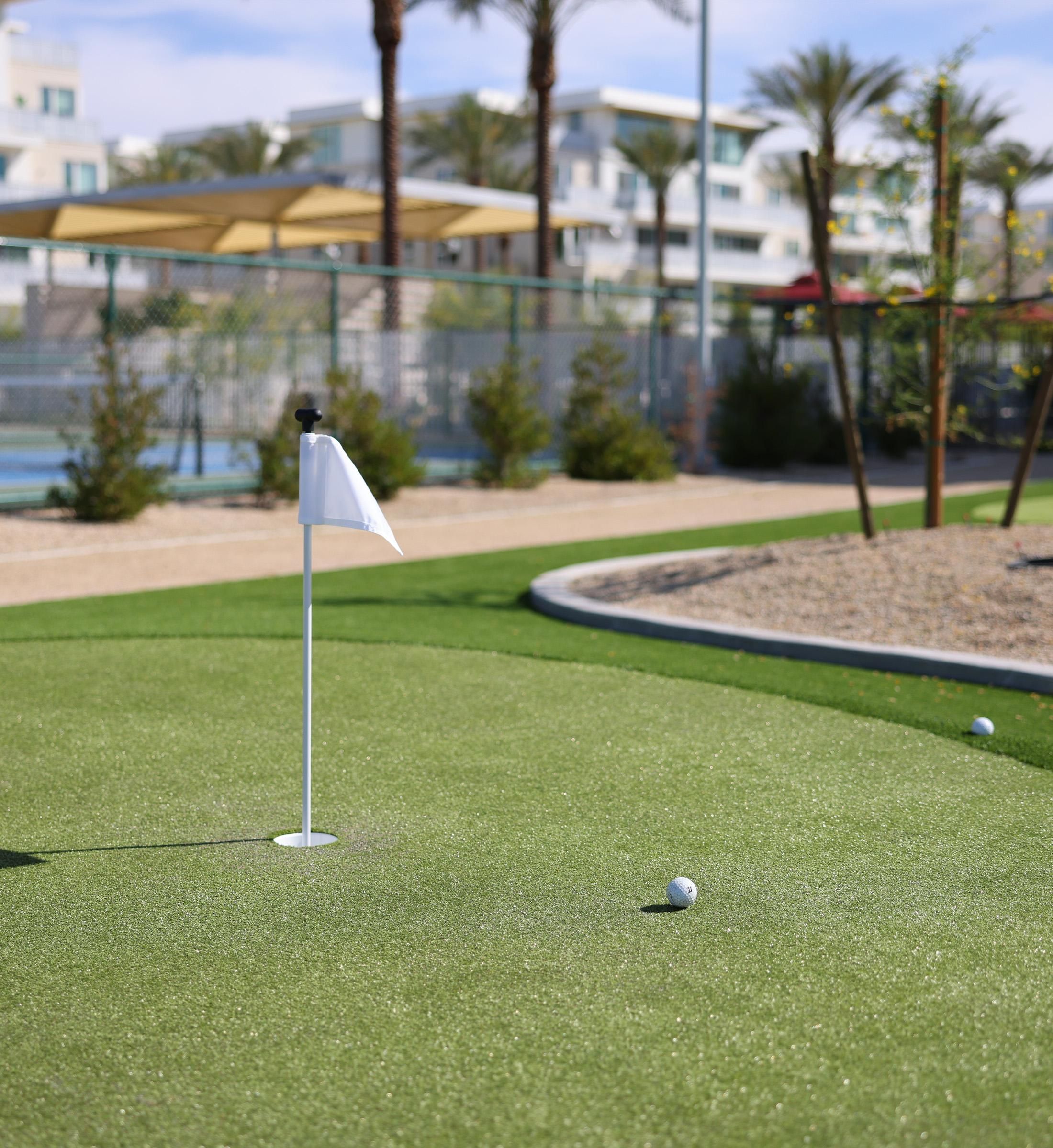 A golf ball near a flag on a putting green, with buildings and palm trees in the background.