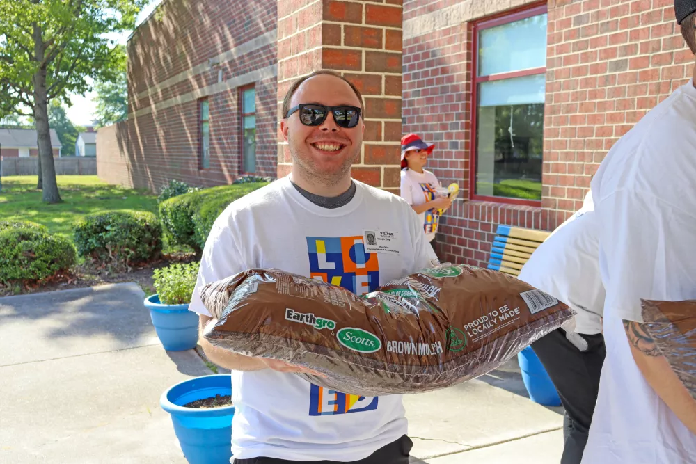 Smiling man in sunglasses holds a large bag of gardening soil outdoors by a brick building with blue planters.