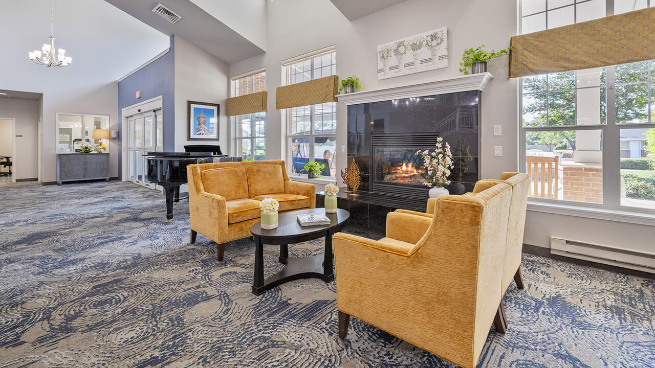 A bright, spacious lounge with patterned carpet, mustard-yellow sofas around a black coffee table, and a fireplace. Large windows let in natural light; potted plants and flowers add a welcoming touch.