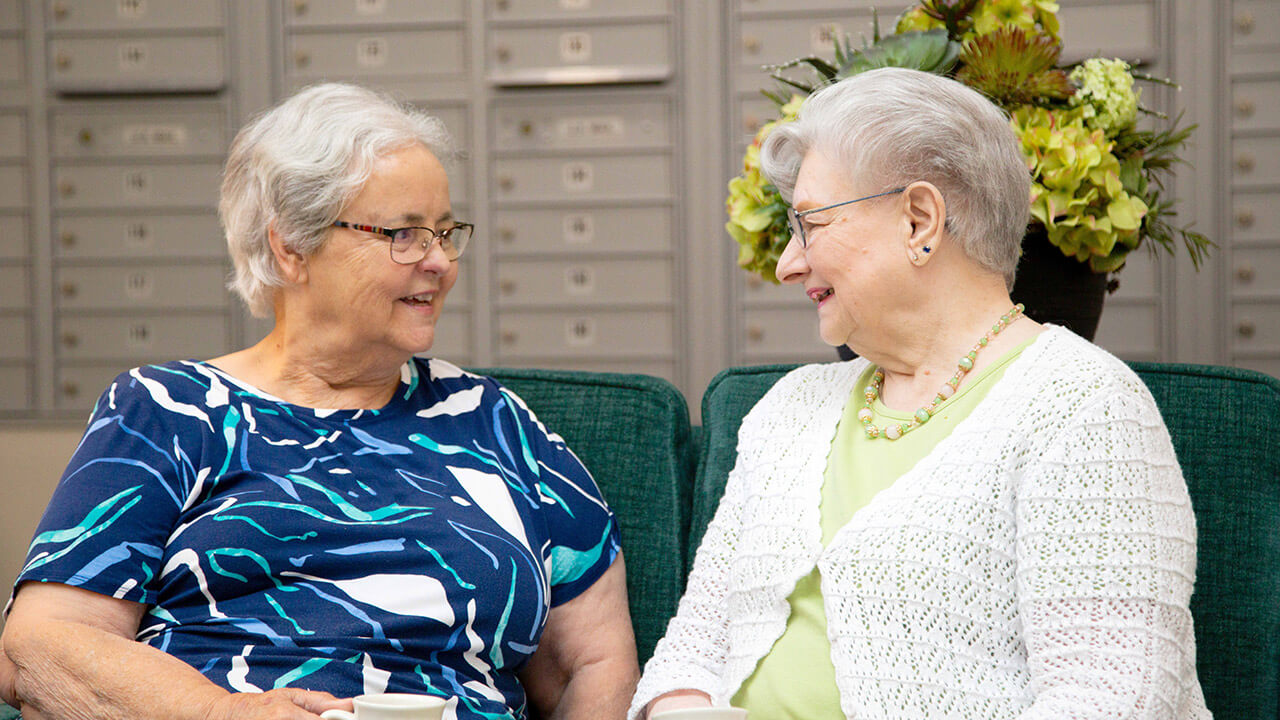 Two elderly women sit on a green couch, smiling and talking. One wears a blue patterned top, the other a white cardigan. Behind them are gray mailboxes and a vase of green flowers.