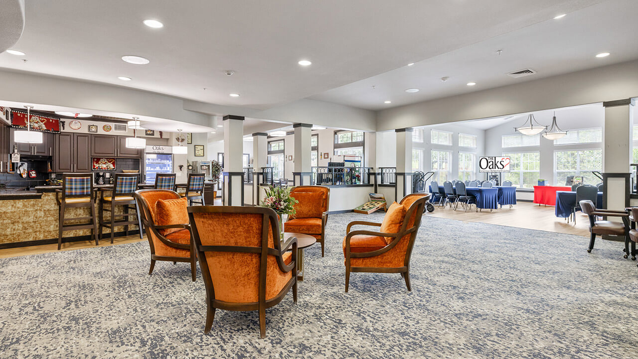 A spacious, well-lit lounge area with four vintage orange chairs around a small table, a bar with stools in the background, and a dining area with tables and chairs by large windows.