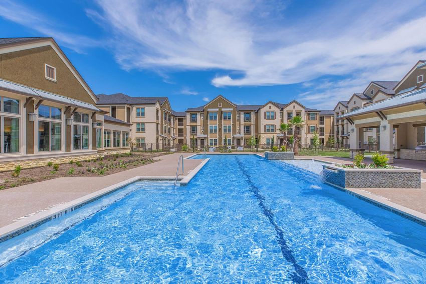 A bright blue swimming pool surrounded by modern apartment buildings. The pool features a shallow area and a landscaped patio with green grass and palm trees. The sky is clear with a few clouds, creating a vibrant outdoor atmosphere.