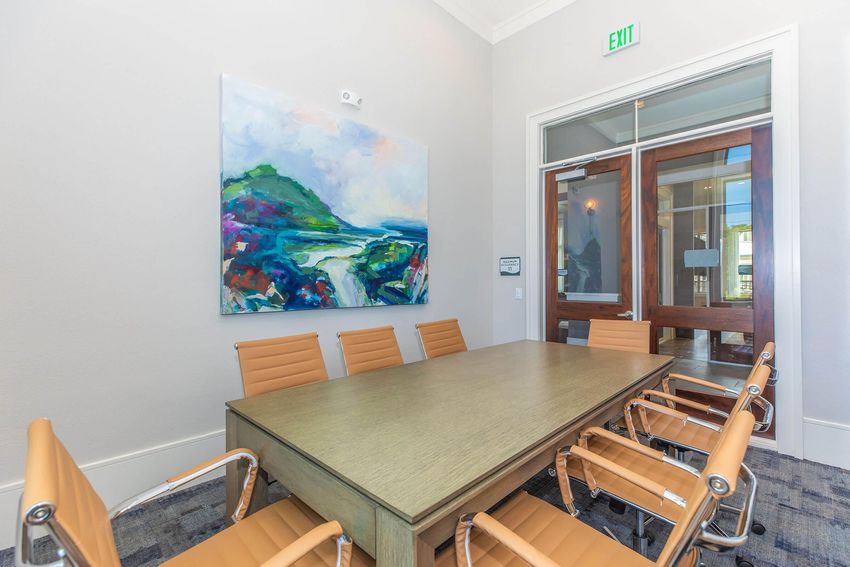 A modern conference room featuring a large wooden table surrounded by orange leather chairs. A colorful abstract painting hangs on the wall, depicting a landscape with hills and a vibrant sky. Glass doors lead to an exterior space, enhancing the room's bright atmosphere.