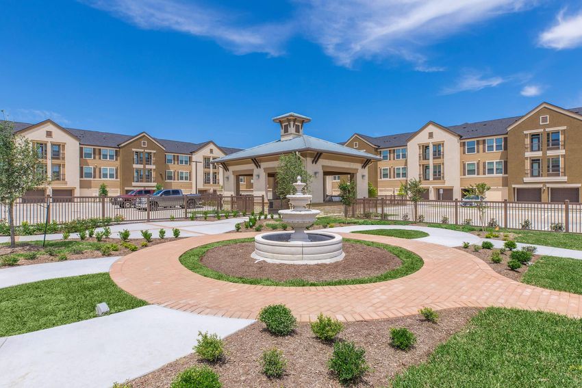 A landscaped courtyard featuring a central fountain surrounded by neatly trimmed shrubs and flower beds. In the background, well-maintained apartment buildings are visible under a bright blue sky with a few clouds. Pathways made of brick and concrete lead through the greenery.