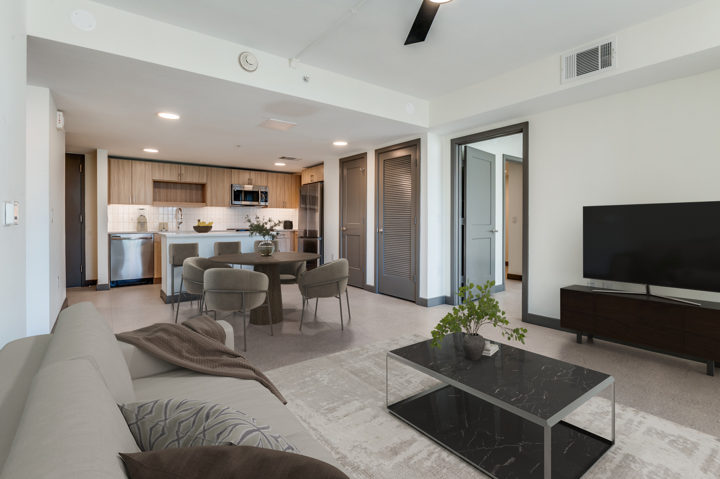Modern open-plan living room and kitchen in Cabana Design District Apartments, featuring neutral decor, a dining table for four, sofa, coffee table, TV stand with large TV. Two doorways lead to other rooms.