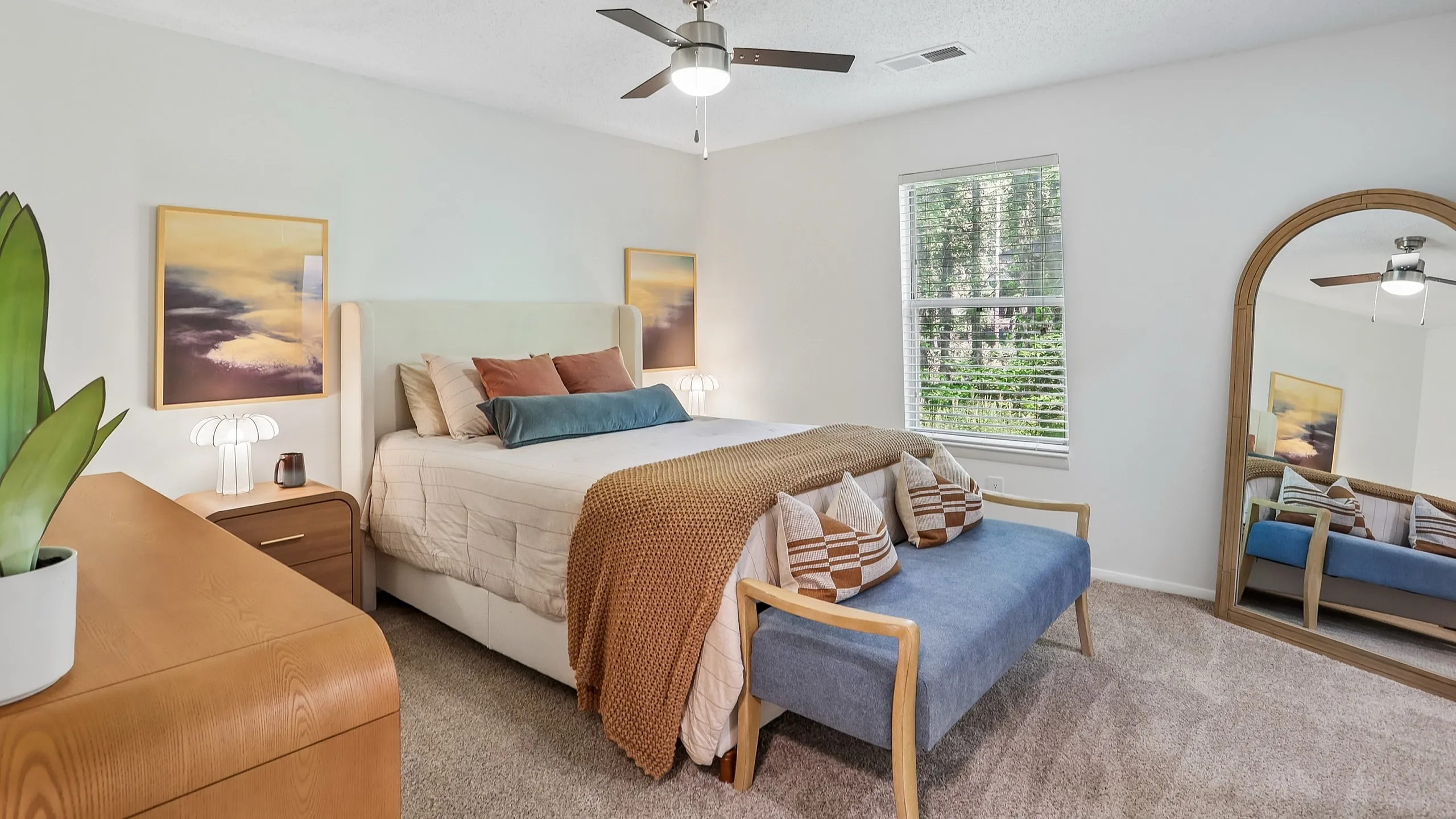 Elegant bedroom with cozy plush bed, vibrant pillows, stylish decor, and a large mirror reflecting natural light from a window with serene outdoor views.