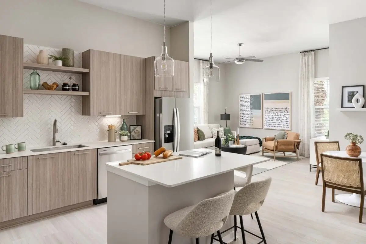 Modern kitchen with light wood cabinets, white countertops, and a small island with two beige barstools. This inviting Windsor Communities space opens into a bright living area with a sofa, armchair, and large windows letting in natural light.