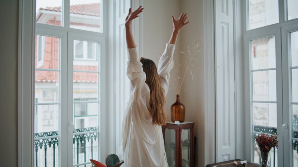 A woman stretching her body while standing by a window, gazing at the beauty of the outdoors