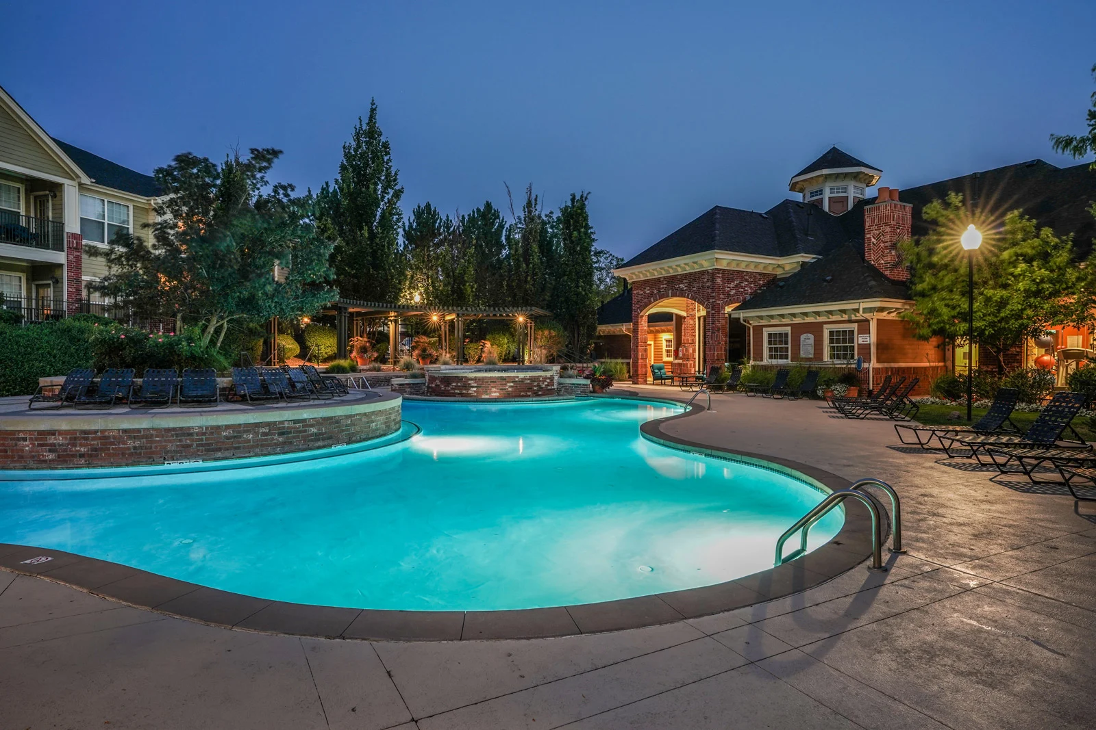 A glowing outdoor swimming pool at dusk at Windsor Meadow Hills, surrounded by lounge chairs, trees, and landscaped gardens, with a brick building and gazebo in the background under a deep blue evening sky.