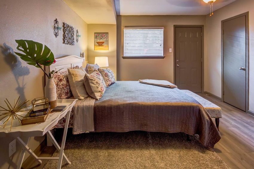 Cozy bedroom with a neatly made bed featuring decorative pillows and a soft throw. A small white bedside table holds a plant and a decorative item. Natural light streams through a window with blinds, and a door is visible on one side. The walls are painted in neutral tones, creating a warm atmosphere.