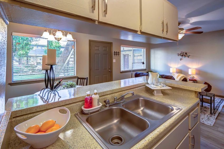 A modern kitchen view featuring a double sink, a bowl of oranges on the countertop, beige cabinets, and a window with natural light. In the background, a living area is visible with a sofa, side tables, and soft lighting, creating a warm and inviting atmosphere.